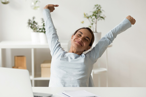 Happy woman stretching in front of desk at work holding arms above head. Female office worker relaxing in cozy chair. Young student having rest at home after finished study. People lifestyle concept