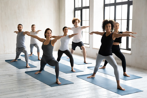 Multinational millennial girls and guys standing barefoot on mats performing Warrior II or Virabhadrasana position. Session led by mixed-race woman trainer showing exercise to people during yoga class