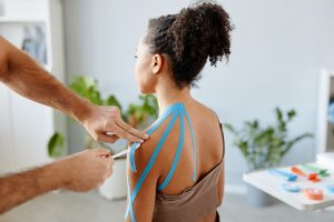 Close up rehabilitation therapist putting k tape on shoulder of young African American woman in physiotherapy clinic