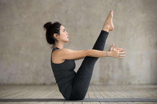 Young yogi woman practicing yoga, doing Paripurna Navasana exercise, boat pose, working out, wearing sportswear, black pants and top, indoor full length, gray wall in yoga studio