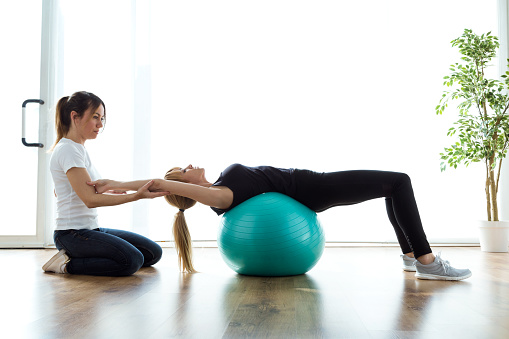 Shot of physiotherapist helping patient to do exercise on fitness ball in physio room.